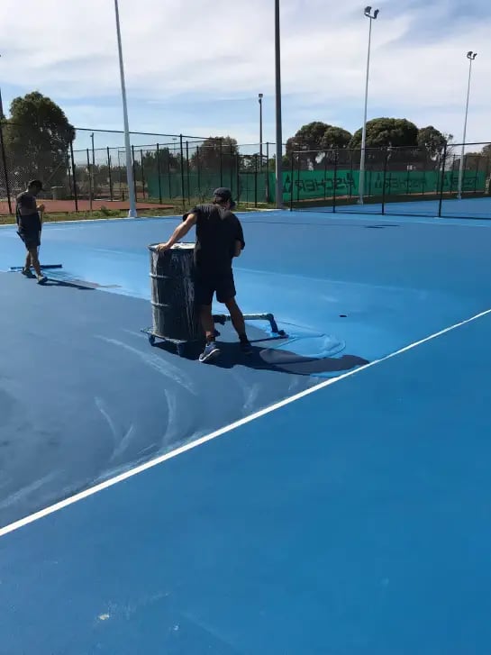 Technician laying blue paint on outdoor tennis court with acrylic surface