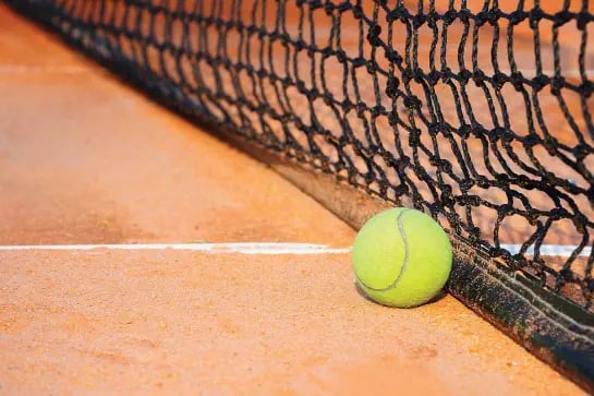 Close up of a tennis ball on a red porous tennis court surface with tennis net behind it